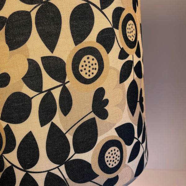 Handcrafted lampshade in graphic bloom fabric with repeating pattern of stylized flowers and leaves set against an off white background, close up, lit.