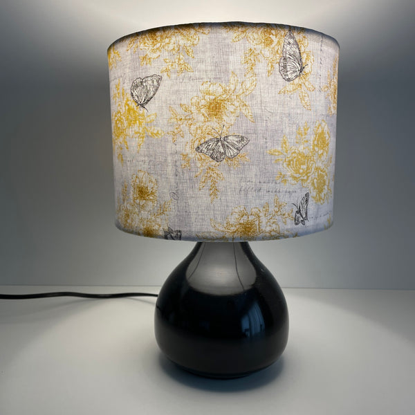 Small drum lampshade in summer romance fabric on small black lamp base, lit, sold by shades at grays