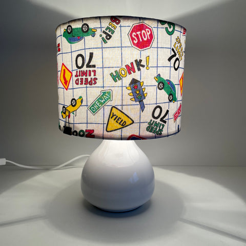 Small fabric hand crafted lamp shade with fun road signs and cars on white background, lit.