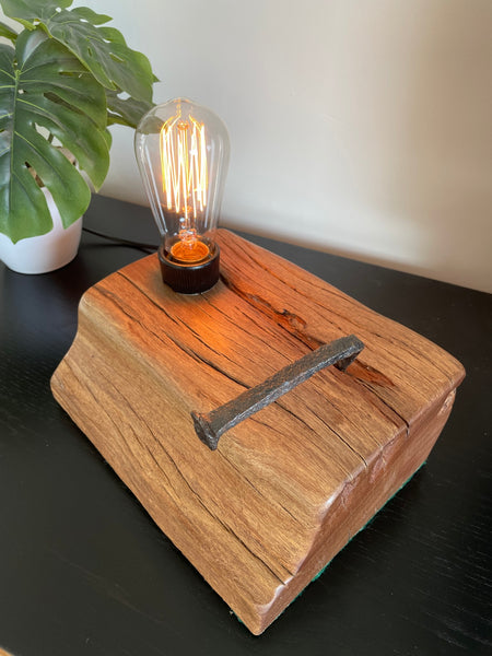 Rustic reclaimed wharf timber table lamp crafted by shades at grays, nz, with original iron nail, lit, angled view of smooth end grain.