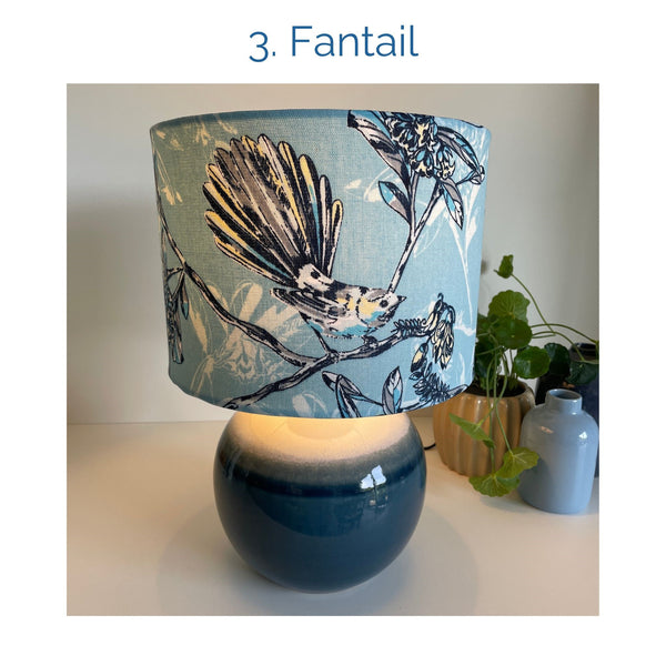 Fantail fabric lampshade on blue ceramic base, lit, by shades at grays.