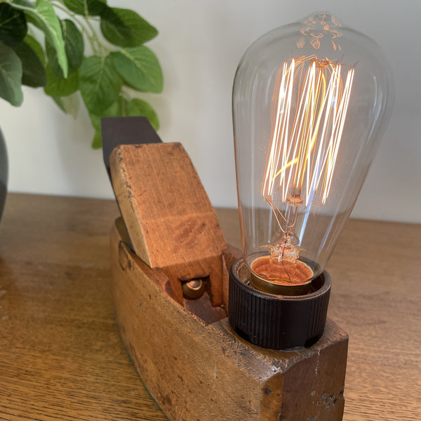 Authentic carpenters' plane upcycled into wood table lamp with edison bulb, lit, crafted by shades at grays, close up of bulb and angled view of plane.