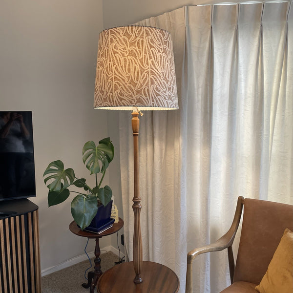 Bespoke tapered lampshade with ihi fabric Pākākā on wooden floor stand, lit.