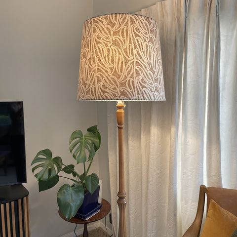 Bespoke tapered lampshade with ihi fabric Pākākā on wooden floor stand, lit.