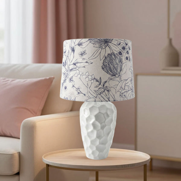 Botanical print lampshade handcrafted by shades at grays, lit on medium white textured ceramic base in living room.