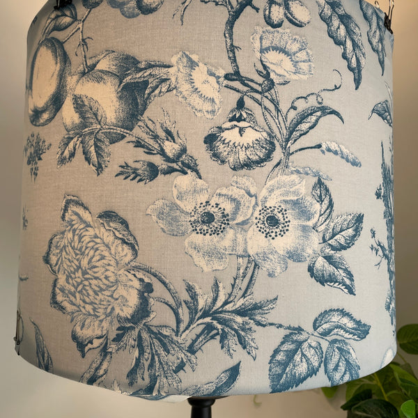 Close up of large drum fabric lampshade with large flowers and fruit etched in steel blue and white on pastel blue background, lit.