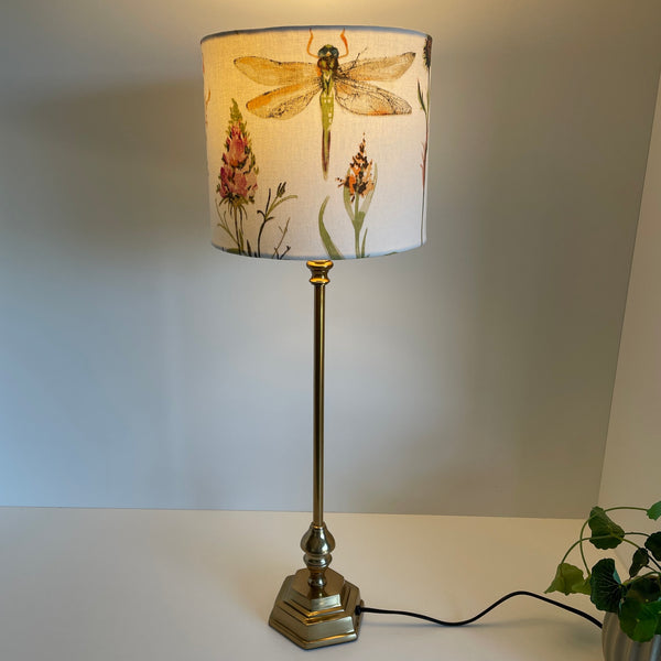 Cream dragonfly fabric on medium cylinder lampshade, lit, on medium high gold base.