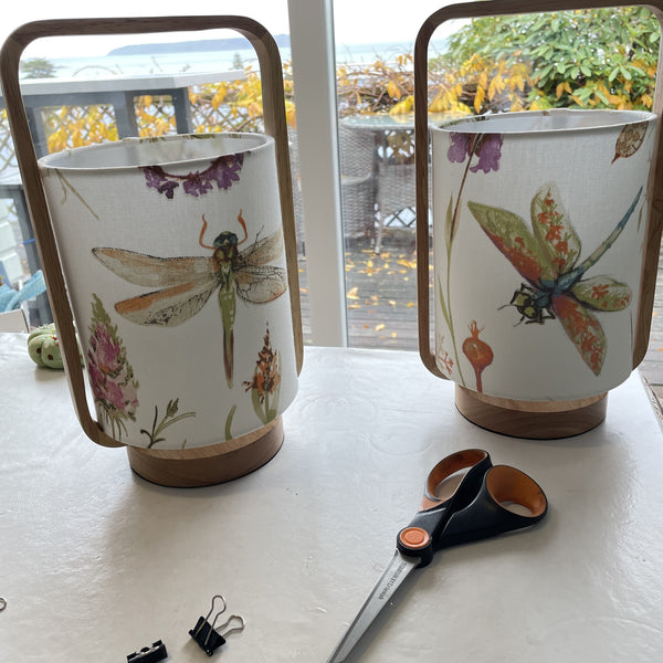 Dragonfly patterened fabric on natural timber table lamps, unlit. Table in foreground and deck view in background.