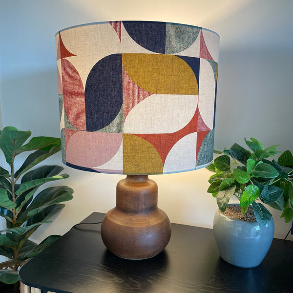 Drum lampshade with Bold and Beautiful fabric on java wood base, lit, on black  cabinet.