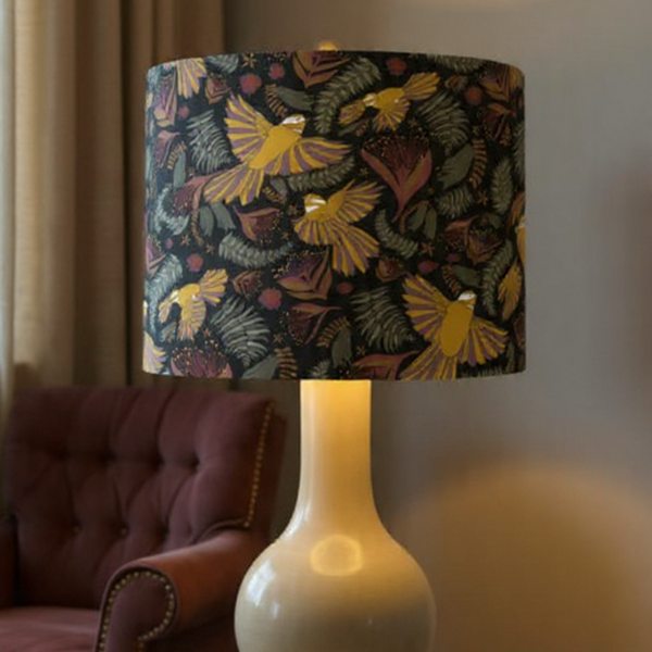 Drum lampshade with Jade Ell's fantail dance on midnight black fabric, handcrafted by shades at grays, on glass base, lit.