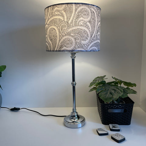 Drum style lamp shade on silver base with grey paisley fabric, lit.