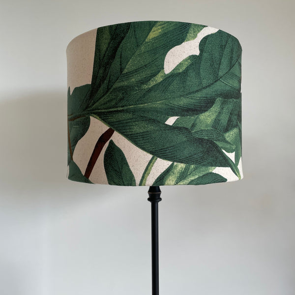 Drum style light shade with rich green leaves and brown stem on textured warm cream background, on black lamp base.