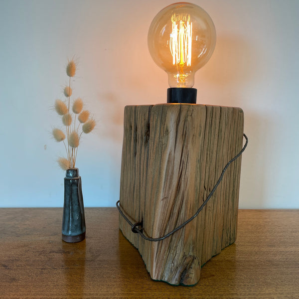 A rustic table lamp made from a natural tōtara fence post with a black metal wire handle and a glowing clear glass Edison bulb on top, set on a wooden tabletop next to a small vase of dried grass against a plain wall.