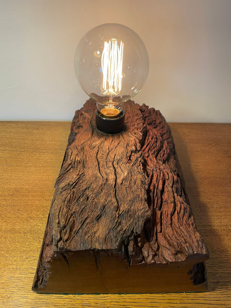 Rustic wood table lamp handcrafted from upcycled jarrah wharf timber, long view of top and smooth end grain with replica edison bulb, lit, by shades at grays.