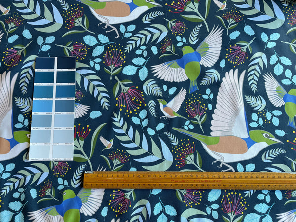 A close-up of Jade Ell's fabric 'Forest Birds, teal' featuring blue and green birds, and light blue leaves on a dark teal background, with a wooden ruler and a small colour swatch card placed on top for scale and colour comparison, by shades at grays.