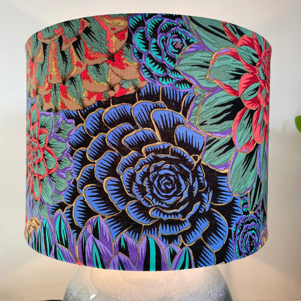 Close up of blue and green flowers on Kaffe Fassett House Leeks Dark fabric on handcrafted lamp shade made in nz by shades at grays, on glazed ceramic base.