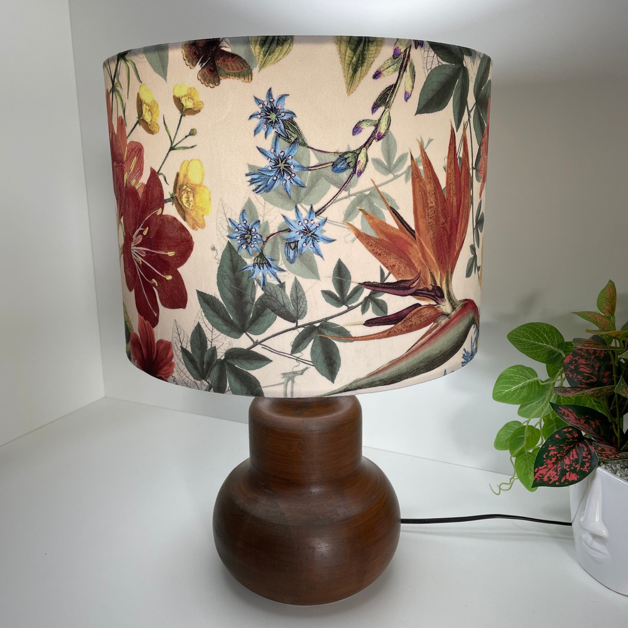 Large drum lampshade handcrafted by shades at grays with velvet fabric showing dainty blue and yellow flowers and bold red and orange blooms on a cream background, lit.