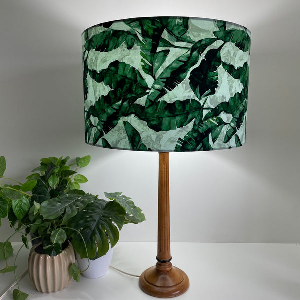 Large barrel bespoke lamp shade with a banana leaves fabric on wooden stand, lit.