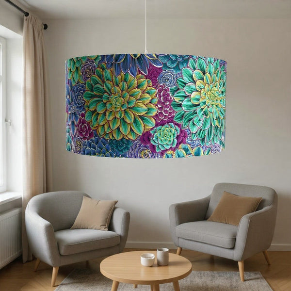 A large barrel pendant light with a vibrant, colorful succulent and floral pattern hangs in a modern living room setting above two gray armchairs and a round wooden coffee table.