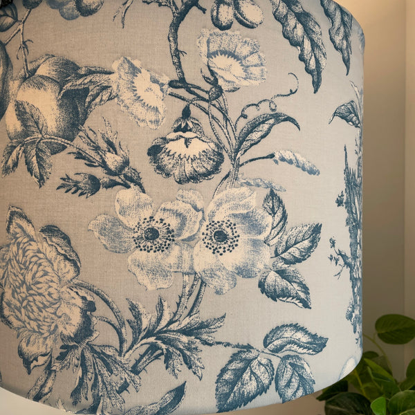 Large drum fabric lampshade with large flowers and fruit etched in steel blue and white on pastel blue background, close up lit.