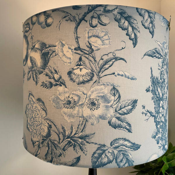 Large drum fabric lampshade with large flowers and fruit etched in steel blue and white on pastel blue background, lit, by shades at grays, nz.
