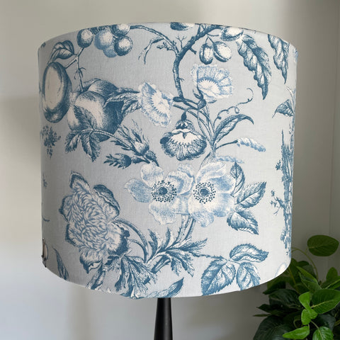 Large drum fabric lampshade with large flowers and fruit etched in steel blue and white on pastel blue background, unlit.