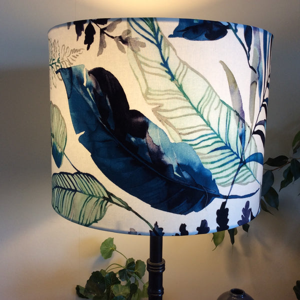 Large drum handcrafted fabric lamp shade with watermark palm fabric, lit. on black stand.