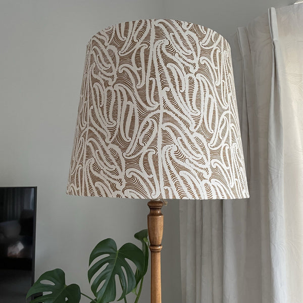 Large tapered lampshade with ihi fabric Pākākā on wooden floor stand, unlit.