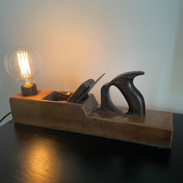 Large vintage carpenters plane upcycled into wood lamp with edison bulb, lit, polished with natural beeswax by shades at grays, front view.