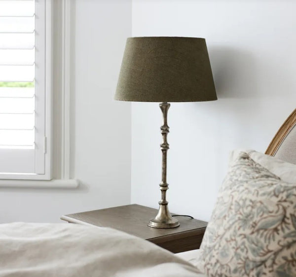 Antique silver candlestick lamp base on a bedside table with an olive tapered shade, a styling example