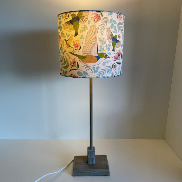 Medium cylinder lampshade with Jade Ell's Tauhou Silvereye Birds fabric, handcrafted by shades at grays, lit on grey base.
