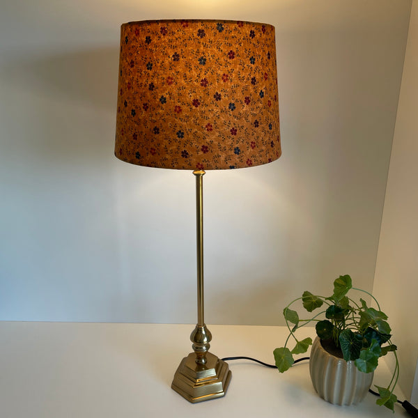 Medium tapered lampshade with cornflowers on brown background, handcrafted by shades at grays, lit on gold octagonal base.