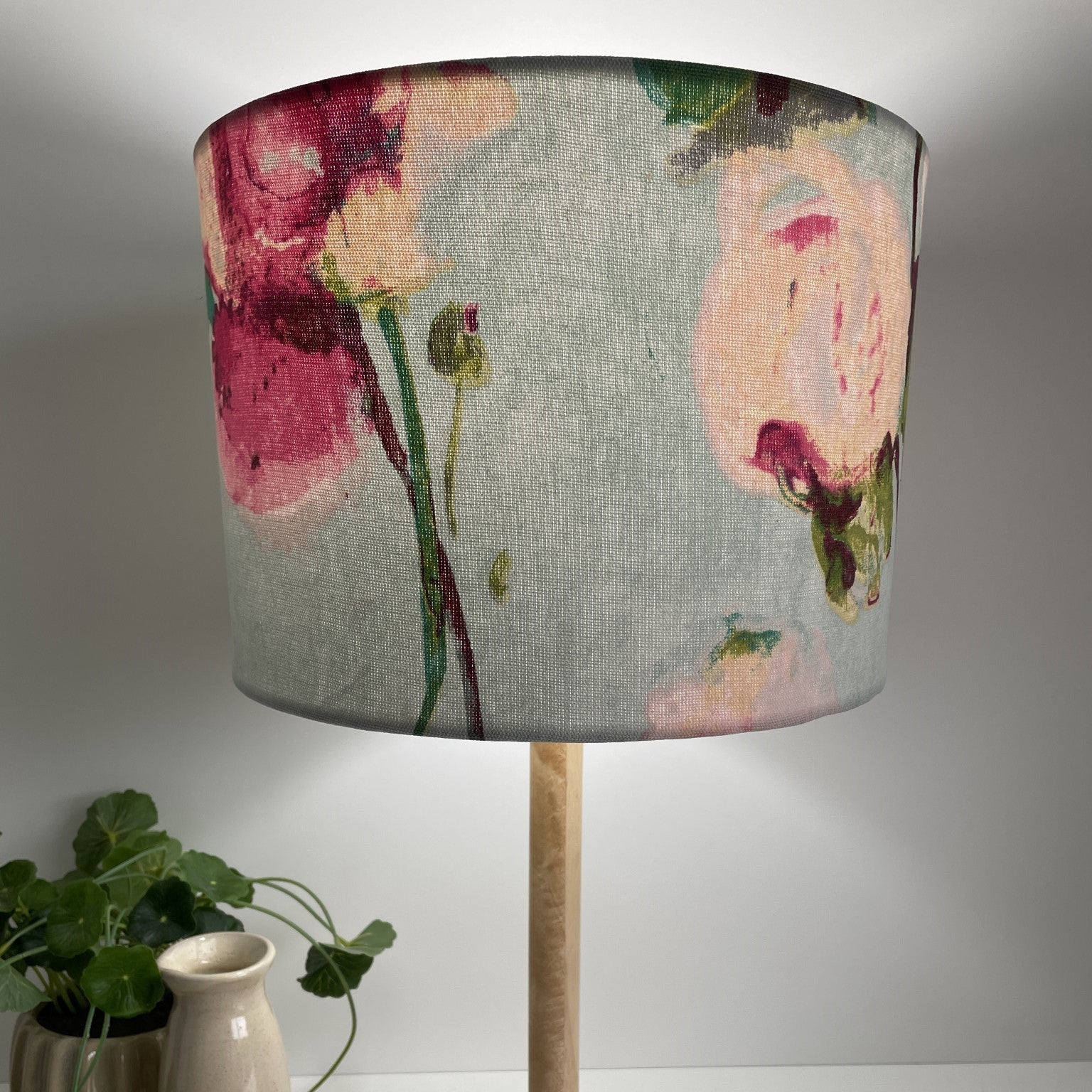 Pink and cream blooms with green stalks on sky blue background, medium drum lampshade, lit.