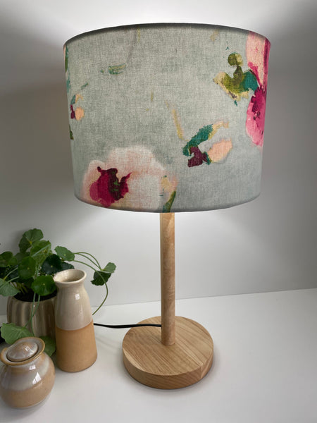 Pink blooms with green stalks on sky blue background, medium drum lampshade, lit, on natural wood lampbase.