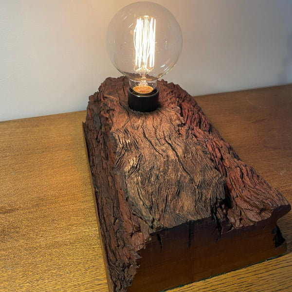 Rustic wood table lamp handcrafted from upcycled jarrah wharf timber, long view of top and sides with replica edison bulb, lit, by shades at grays.