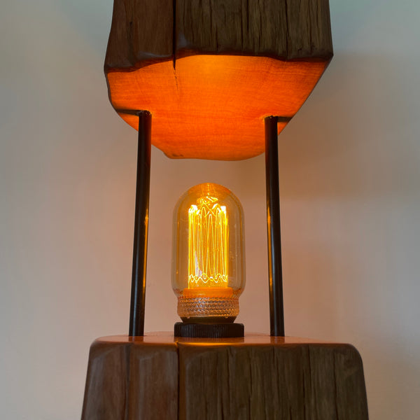 Reclaimed wood table lamp crafted with a split totara post and black metal rods, glow of  bulb.