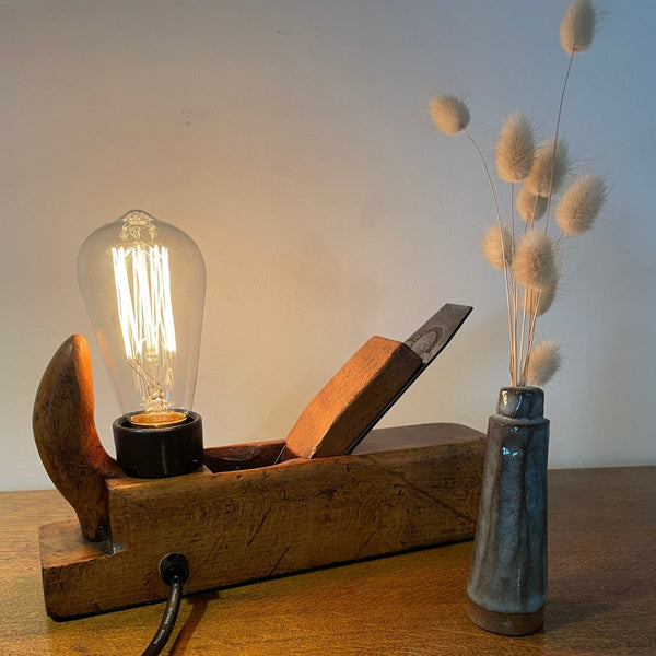 Wood table lamp crafted from authentic carpenters plane, with replica teardrop bulb, lit, back view with black lead by shades at grays