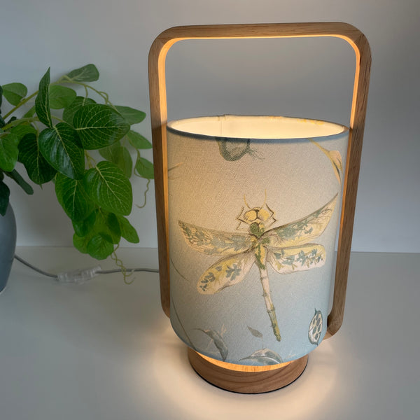 Scandi style table lamp with blue dragonfly fabric, lit, by shades at grays.