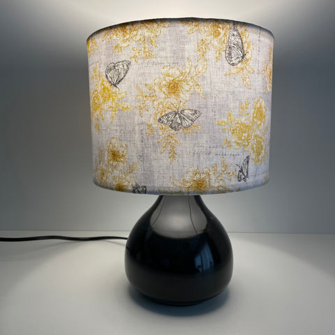 Small drum lampshade in summer romance fabric on small black lamp base, lit, sold by shades at grays