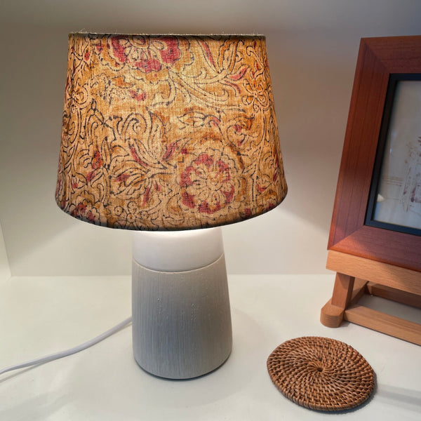 Small tapered lampshade handcrafted by shades at grays with mustard block print fabric, lit on small beige table lamp.