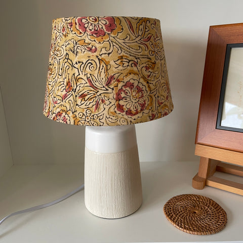 Small tapered lampshade handcrafted by shades at grays with mustard block print fabric,unlit on small beige table lamp.