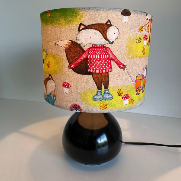 Small drum lampshade in fox fabric, lit, on small black base, handcrafted by shades at grays.