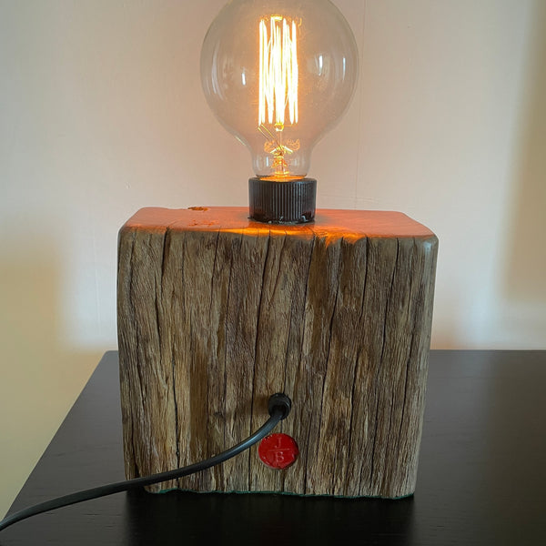 Smalll wood lamp crafted from recycled wharf timber with original bolt and edison bulb, lit, back view, by shades at grays.