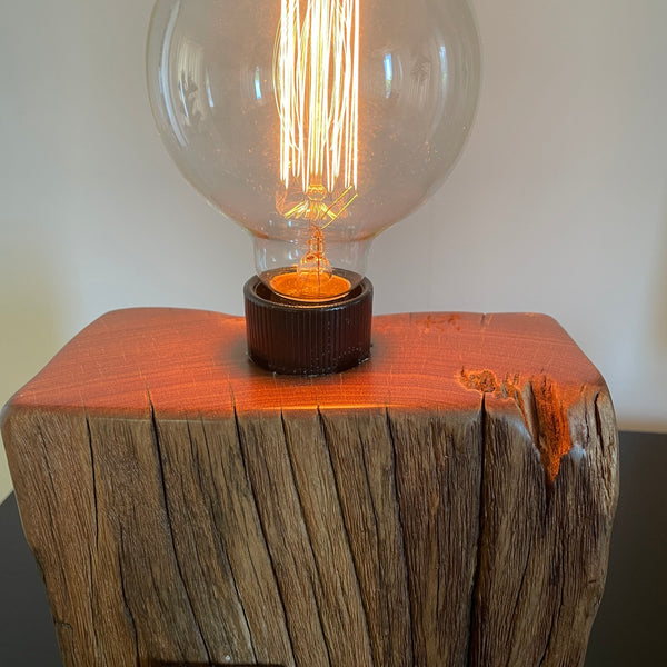 Smalll wood lamp crafted from recycled wharf timber with original bolt and edison bulb, lit, close up of polished top and erosion lines, by shades at grays.