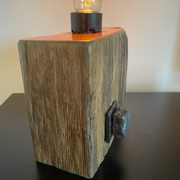 Smalll wood lamp crafted from recycled wharf timber with original bolt and edison bulb, lit, left side, by shades at grays.