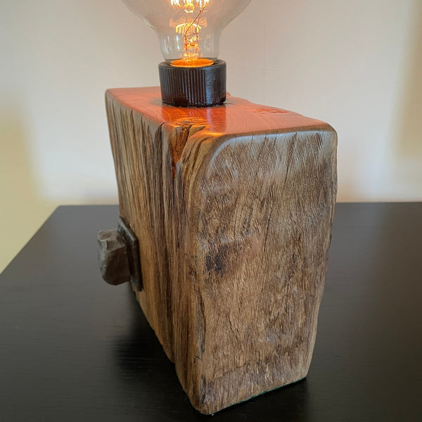 Smalll wood lamp crafted from recycled wharf timber with original bolt and edison bulb, lit, right side, by shades at grays.