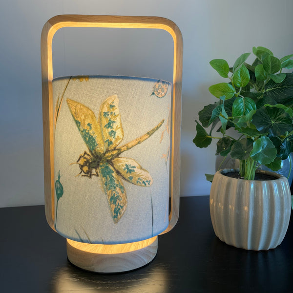 Table lamp with natural wood frame and blue dragonfly fabric, lit, by shades at grays.