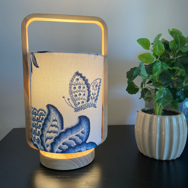 Table lamp with plain wood frame and blue embroidered butterfly and flower on white background, lit.