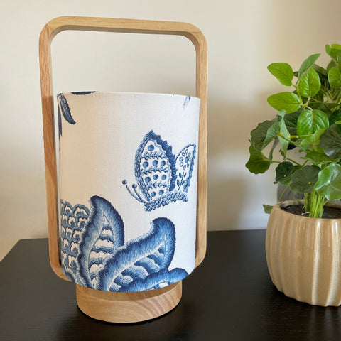 Table lamp with plain wood frame and blue embroidered butterfly and flower on white background, unlit.