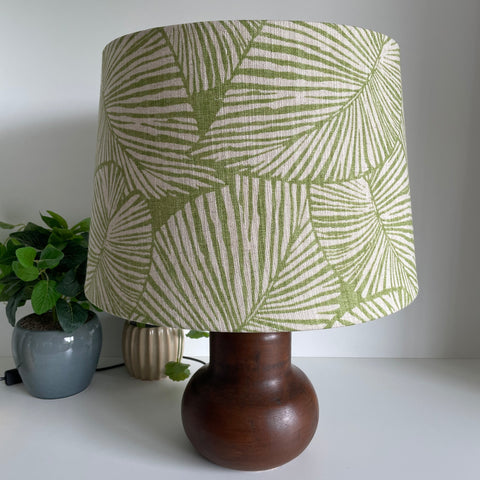 Tapered 46 lampshade handcrafted by shades at grays with large pattern of green palms, unlit on brown base.